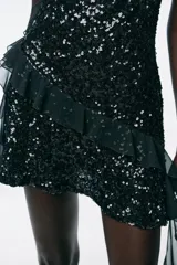 Vestido corto de escote fluido y tirantes finos, completamente cubierto de lentejuelas negras. Presenta detalles de volantes en tejido combinado que recorren el lateral y el frente de la prenda.