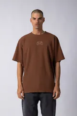 Remera de corte oversize color marrón, con cuello redondo y mangas cortas con hombros caídos. Presenta un bordado minimalista de un joystick en el centro del pecho y una etiqueta aplicada en la parte superior de la espalda.