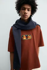 Camiseta de corte relajado color marrón, con cuello redondo y manga corta. Presenta un parche central con diseño gráfico colorido y abstracto.