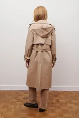 Trench coat color beige con cuello de solapa, cierre frontal abotonado, bolsillos laterales y mangas largas con cintas ajustables. Incluye capucha desmontable y forro interior rayado. La tela exterior cuenta con protección repelente a la lluvia y manchas.