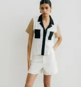 Camisa de manga corta con diseño color block, cuerpo principal blanco con detalles en negro (cuello y tapeta de botones) y mangas en color beige/tostado. Presenta dos bolsillos cuadrados negros en el frente.