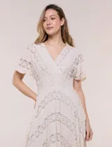 Vestido midi blanco de estilo romántico confeccionado en poliéster con elastano, con detalles de encaje floral. Cuenta con escote cruzado en V, mangas cortas acampanadas, cintura elastizada y falda evasé con terminación asimétrica en picos.