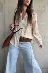 Camisa de manga larga color beige con detalles de encaje y calados verticales en el frente y las mangas, cuello en V y cierre frontal con botones.