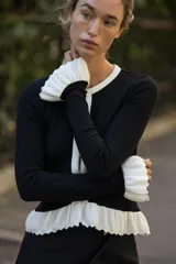 Chaqueta de punto acanalado en color negro con detalles en blanco en cuello, tapeta de botones, bolsillos y volados en el ruedo y puños. Presenta cierre frontal con botones dorados.