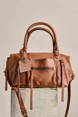 Cartera de cuero color tostado con acabado gastado, doble asa de mano, correa ajustable y desmontable, y múltiples bolsillos exteriores con cierre y tiradores de cuero.