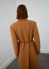 Abrigo de lana color marrón camel, de corte recto y largo midi, con solapas de muesca, bolsillos frontales con solapa y manga larga. Incluye un cinturón fino de cuero oscuro para entallar la cintura.