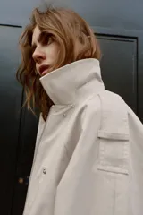 Trench corta de color beige, con cuello solapa, mangas largas con trabillas en los hombros, bolsillos delanteros de vivo y cierre frontal cruzado con botones a presión.