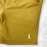 Bermuda deportiva color mostaza, con cintura elástica ajustable con cordón y bolsillos laterales. Presenta un logo pequeño bordado en la pierna derecha.