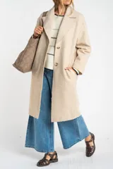 Tapado largo de corte oversize en color beige, con solapas clásicas y cierre frontal con botones. Presenta un diseño minimalista y elegante, ideal para un look sofisticado.