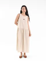Vestido largo sin mangas de color beige, con calce amplio y relajado, escote redondo y falda fruncida. Cuenta con detalles de bordado a contratono en las sisas y logo frontal.