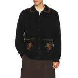 Buzo tipo cárdigan de punto color negro, con cierre frontal de botones, cuello tipo polo y dos bolsillos frontales. Presenta detalles de costura decorativa en color contrastante en el cuello y los bolsillos, además de un bordado floral en cada bolsillo.