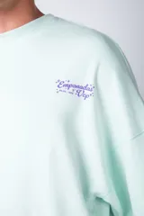 Buzo oversize de cuello redondo en color verde menta, confeccionado en tela rústica. Presenta un pequeño estampado en el pecho y una serie de estampados verticales a lo largo de la manga izquierda.