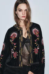 Chaqueta abierta de terciopelo negro con cuello redondo y mangas largas acampanadas. Presenta bordados florales en tonos rosados y verdes, con aplicaciones de abalorios y detalles dorados en el cuello y la parte frontal.