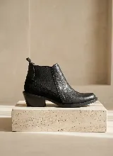 Bota tipo Chelsea de cuero negro con paneles laterales elásticos y taco bajo cuadrado. Presenta una combinación de texturas en el cuero y una presilla trasera para facilitar el calce.