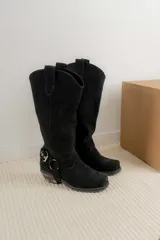 Bota estilo tejana de caña alta, confeccionada en cuero marrón con acabado texturizado. Presenta punta cuadrada, taco bajo y una correa con hebilla metálica ajustable y estribo desmontable en la zona del tobillo.