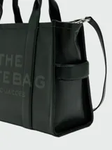Bolso tipo tote de cuero negro, modelo The Leather Medium Tote Bag de Marc Jacobs. Presenta doble asa superior, correa ajustable para el hombro y el texto grabado "THE TOTE BAG MARC JACOBS" en el frente.