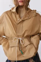 Cazadora tipo anorak color beige, con capucha ajustable, manga larga y bolsillo frontal de plastrón con solapa. Cuenta con cordones elásticos ajustables en la cintura y el bajo, y cierre frontal con botones a presión.