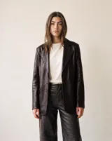 Blazer de cuero marrón oscuro, de corte sastre con solapas de muesca, cierre frontal de un botón y bolsillos con solapa en los laterales.