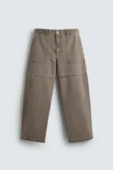 Pantalón de corte relajado en color marrón con efecto lavado. Presenta bolsillos frontales tipo cargo, cierre con cremallera y botón, y una línea horizontal de tachas metálicas a tono en la parte superior de las piernas.