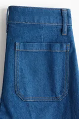 Pantalón de jean de tiro alto, color azul medio, con pierna ancha y corte ajustado en la cadera. Presenta bolsillos de parche tanto en la parte delantera como en la trasera.