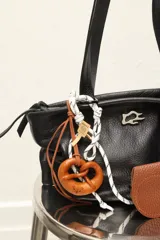 Charm para bolso con forma de pretzel trenzado en cuero color caramelo, sujeto con cordón blanco y negro trenzado. Incluye un pequeño dije rectangular de madera con grabado.