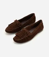 Mocasines de mujer Satinato confeccionados en cuero de gamuza marrón, con flecos, moño decorativo y costuras visibles en la capellada. Presentan punta redondeada y suela baja.