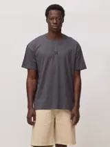 Remera de corte oversize color gris oscuro, con cuello redondo y pequeño detalle bordado en forma de cruz en el centro del pecho.