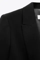 Blazer negro de corte recto, con cuello solapa y hombreras. Las mangas son tres cuartos con vuelta asegurada por un botón dorado. Posee bolsillos delanteros con solapa.