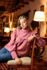 Blusa rosa de algodón con cuello alto y mangas largas abullonadas. Presenta detalles de cinta negra en el cuello y los puños, hombros con diseño princesa y cierre de lágrima con botón forrado en la parte posterior del cuello.