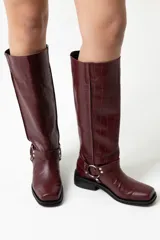 Botas de cuero croco color rojo oscuro, de caña alta (38 cm), con taco bajo de 3 cm y punta cuadrada. Presentan un arnés decorativo con hebilla metálica alrededor del tobillo.