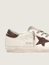 Zapatillas Golden Goose modelo Super-Star, color blanco con detalles en marrón con glitter. Confeccionadas en cuero vacuno, con estrella lateral y contrafuerte de talón brillantes, punta y lengüeta de gamuza, y efecto envejecido en la suela.