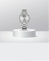 Reloj analógico de pulsera con caja redonda de metal plateado, bisel decorado con cristales y malla de eslabones metálicos. Cuenta con movimiento de cuarzo japonés y cierre desplegable con botón de seguridad.