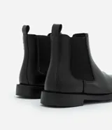 Bota tipo Chelsea de caña media, color negro, con paneles laterales elásticos y suela de goma. Diseño de punta redonda y estilo minimalista.