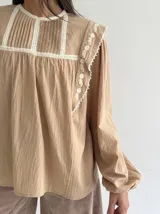 Blusa color beige de algodón con cuello redondo, mangas largas con puños elásticos y detalles de puntilla y bordados en el frente y hombros.