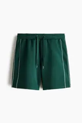 Short de corte relajado de color verde oscuro, con cintura elástica y cordón ajustable. Cuenta con ribetes de cinta blanca a lo largo de los laterales y bolsillos frontales.