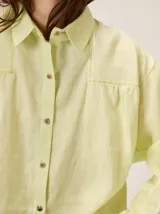Camisa de lino color verde lima, con cuello clásico, cierre frontal de botones y mangas largas con puños. Presenta detalles de pespuntes en zigzag en la zona de los hombros y canesú.