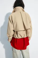 Trench corto de cuello solapa y escote en pico, color beige, con mangas largas terminadas en trabillas con botones. Cuenta con bolsillos laterales con solapa, cierre frontal cruzado con botón y bajo abullonado con elástico interior.