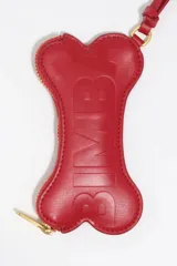 Monedero pequeño de cuero reconstituido color rojo brillante, con forma de hueso de perro. Posee cierre de cremallera dorado y una correa corta de cuero para sujetar.