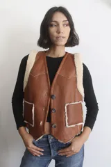 Chaleco de cuero genuino color marrón con interior y bordes de corderito sintético. Presenta cierre frontal con botones, bolsillos aplicados y diseño estilo western.