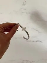 Brazalete tipo esclava rígida abierta, fabricada en acero quirúrgico, con un detalle de cruz integrada en la estructura.