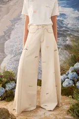 Jean wide leg color crudo, con tiro alto, bolsillos delanteros y traseros, costuras pespunteadas a contratono y pequeños bordados decorativos dispersos.