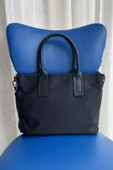 Cartera tipo shopper color negro, de diseño estructurado con doble asa de mano y cierre superior. Presenta sutil grabado de la marca en la parte inferior frontal.