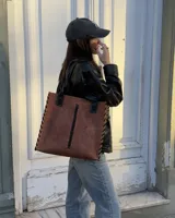 Cartera tipo tote de cuero marrón con diseño estructurado. Presenta detalles de trenzado lateral y central en color negro, con remaches metálicos decorativos. Incluye asas de hombro fijas y el logo de la marca grabado en la parte superior.