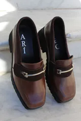 Mocasín de cuero vacuno color bordó con suela track negra de goma. Presenta un diseño de punta cuadrada con un herraje metálico plateado sobre una banda de gamuza en la capellada.