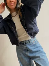 Chaqueta corta de efecto gamuza en color azul marino, con cierre frontal y solapa decorada con una hilera de aros metálicos. Presenta mangas tipo globo, ajuste lateral en la cintura y forro interior.