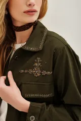 Campera estilo militar o safari color verde oliva, confeccionada en algodón. Presenta cuello solapa y bolsillos frontales con tapa. Destaca por sus apliques de pedrería o bordados en tonos marrones y dorados en el cuello y sobre los bolsillos.