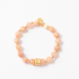 Pulsera elástica compuesta por cuentas de piedra natural en tono rosado, intercaladas con pequeñas esferas doradas. Presenta un detalle central de tres anillos dorados y un dije colgante con forma de moneda con la inscripción MMA.