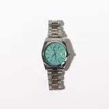 Reloj de pulsera plateado con esfera color turquesa y correa de eslabones metálicos.