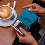 Billetera tarjetero automática con estructura de aluminio y revestimiento de cuero bicolor en tonos verde oliva y beige. Cuenta con un cierre de broche a presión y el logo de la marca grabado en la parte inferior.