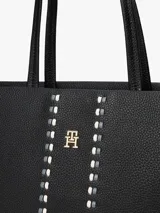 Cartera tipo tote de Tommy Hilfiger en color negro, confeccionada en poliuretano con textura granulada. Presenta doble asa superior, cierre principal y un diseño central decorativo con tachas metálicas y el logo monograma TH en metal dorado.